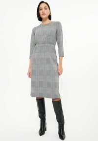 Grau kariertes Kleid mit taillierter Taille, Dreiviertelärmeln und knielangem Saum; getragen mit schwarzen, knielangen Stöckelschuhen.