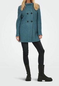 Cappotto doppiopetto color teal con grandi bottoni neri, tasche laterali e vestibilità dritta, abbinato a pantaloni neri attillati e stivali verde scuro.