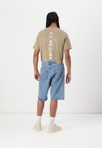 T-shirt graphique beige avec un motif de lune au dos, associé à un short en denim bleu clair. Le modèle porte des chaussettes blanches et des baskets beiges.