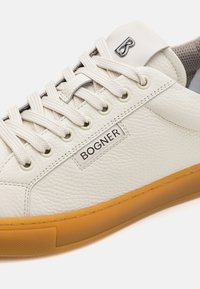 Sneaker bianca in pelle con superficie testurizzata, dotata di suola in gomma con un bordo marrone chiaro e dettaglio del marchio sulla linguetta.