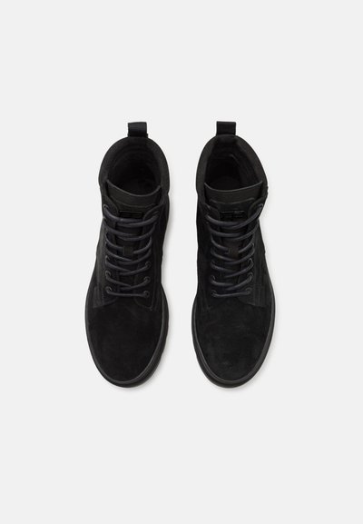 Bottines noires en daim, avec des panneaux texturés, des lacets ronds et une tirette au talon pour un enfilage facile. Semelle plate en caoutchouc.