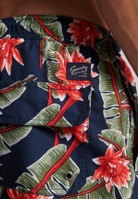 Costumini da bagno corti in blu navy con un motivo floreale che presenta fiori rossi e foglie verdi. Include una tasca e un'etichetta cucita.
