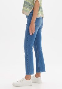Vaqueros de mezclilla azul de corte acampanado con un diseño de tiro medio, combinados con zapatillas blancas. La tela tiene una textura suave y un estilo clásico de cinco bolsillos.