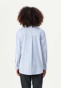 Stripet blå og hvit langermet skjorte med en bakfylling, knappedempere og en buet sømdesign. Laget av et lett, vevd stoff.
