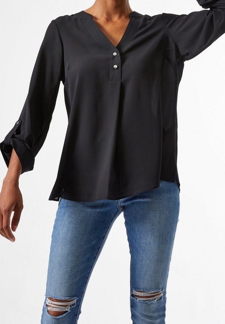 Blusa negra, ligera, con escote en V, tapeta de tres botones y mangas largas que se pueden arremangar. Llevada con jeans azules desgastados que presentan rotos en las rodillas.