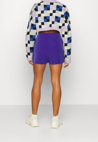 Korthalsad tröja med ett flerfärgat rutmönster; lila shorts; vita sneakers; synlig bakvy och bendetalj.