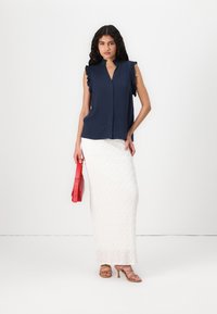 Blusa senza maniche blu navy con dettagli di volant, abbinata a una gonna maxi bianca a trama e una borsa rossa. Modello che indossa sandali beige con cinturino.
