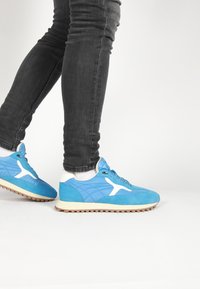 Blue suede sneakers with white accents, lace-up design, textured surface, and rubber sole. Worn with dark slim-fit jeans.