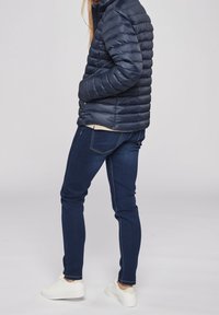Granatowa puchowa kurtka z poziomym przeszyciem, jasną podszewką i bocznymi kieszeniami, zestawiona z ciemnoniebieskimi dopasowanymi jeansami i białymi trampkami.