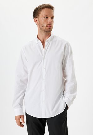 Camisa blanca de botón con mangas largas, textura suave, dobladillo redondeado y cuello abotonado. Llevada con pantalones negros, presentando un ajuste entallado.