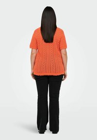 Oranje gebreide top met korte mouwen, voorzien van een gestructureerd patroon en een gegolfde zoom, gecombineerd met zwarte flare jeans. Van achter bekeken.