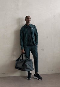 Mees tumehallis Nike sportsikotis hoiab halli spordikotid, seistes kerge tekstuuriga seina vastu siledal põrandal.