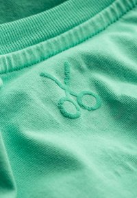 Lichtgroene katoenstof met een gestructureerd oppervlak, met een borduursel van een schaarlogo in een lichtere tint. Elastische tailleband zichtbaar.