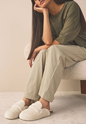 Femme assise sur une chaise, le menton reposant sur la main, portant une chemise verte, un pantalon à carreaux et des chaussons blancs duveteux.