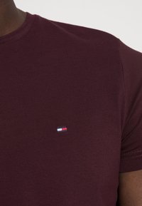 Burgunderrød kortærmet t-shirt lavet af glat stof med et lille broderet logo med røde, hvide og blå elementer på brystet.