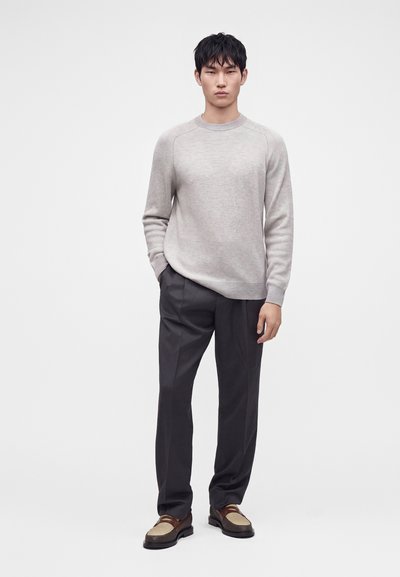 Homme portant un pull gris clair, un pantalon sombre et des mocassins marron et beige, debout avec une main dans la poche sur fond uni.