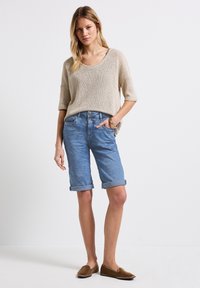 Suéter de punto color beige claro con mangas cortas, combinado con shorts de mezclilla azul claro doblados al final. Calzado marrón sin cordones.