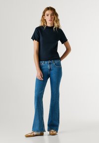 Μαύρο κοντομάνικο τοπ με ριμπ ύφανση και κυματιστά τελειώματα, σε συνδυασμό με ανοιχτό μπλε τζιν παντελόνια με ελαφρύ καμπάνα κόψιμο. Το μοντέλο φοράει καφέ μαλακά παπούτσια με λευκές λεπτομέρειες.