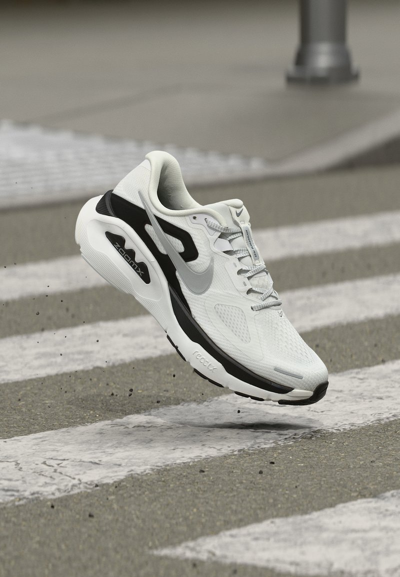 Άσπρο και μαύρο παπούτσι τρεξίματος Nike ZoomX που αιωρείται πάνω από μια διάβαση πεζών, σηκώνοντας μικρά κομμάτια χαλικιού.