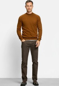 Brauner gestrickter Pullover mit rundem Ausschnitt, getragen über einem weißen Hemd, kombiniert mit dunkelbraunen Jeans und schwarzen klobigen Schuhen.