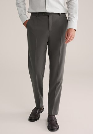 Pantalons gris sur mesure avec une texture lisse, devant plat et deux poches latérales. Accompagnés de chaussures à enfiler sombres, dotées d'une finition en cuir.