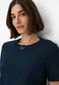 Marineblaue Kurzarm-T-Shirt mit rundem Halsausschnitt, getragen mit einer silbernen Halskette, die zwei miteinander verbundene Ringe zeigt. Glatte Stoffstruktur.
