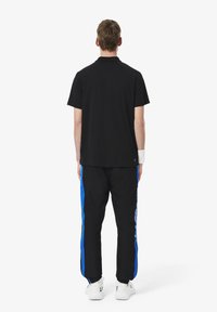 Zwarte polo met korte mouwen, gecombineerd met zwarte broek met blauwe zijstrepen en wit logo. Model draagt witte sneakers.