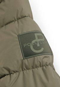 Olivgrön pufferjacka vid axeln med quiltad stoppning, med en rektangulär patch som visar texten “POLO CLUB” i mörkgrönt.