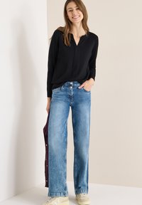 Černá halenka s dlouhým rukávem v kombinaci se světle modrými džíny s širokými nohavicemi. Džíny mají opotřebovaný vzhled a jsou doplněny béžovými teniskami.