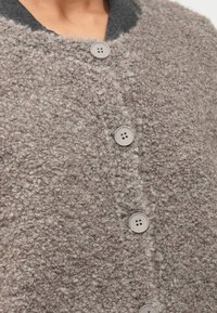 Cardigan texturat de culoare maronie, realizat dintr-un material puțin pufos, asemănător lânii; are cinci butoane rotunde, de culoare deschisă și un guler contrastant de culoare gri închis.