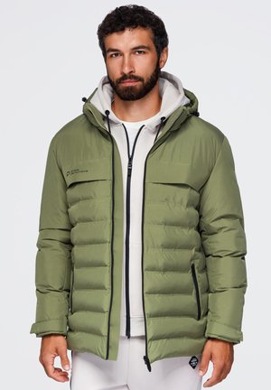 Homme portant une veste matelassée vert olive à capuche par-dessus un sweat à capuche blanc zippé et un pantalon blanc sur un fond uni.