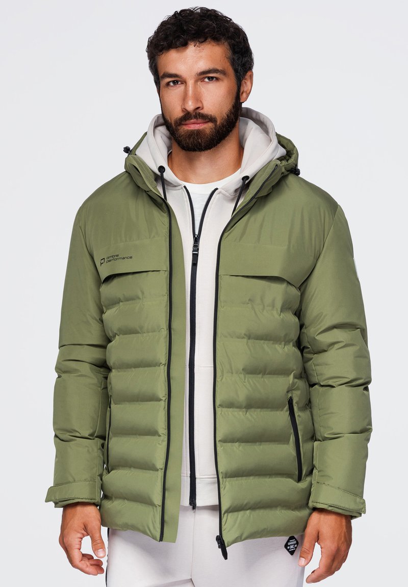 Homme portant une veste matelassée vert olive à capuche par-dessus un sweat à capuche blanc zippé et un pantalon blanc sur un fond uni.