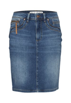 Personne portant une jupe en jean bleu et un haut blanc, avec un détail de tirette marron et des bagues argentées à la main gauche, debout devant un fond uni.