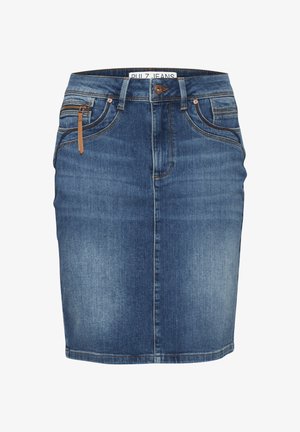 Personne portant une jupe en jean bleu et un haut blanc, avec un détail de tirette marron et des bagues argentées à la main gauche, debout devant un fond uni.