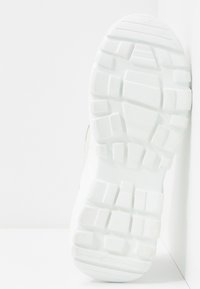 Semelle d'une chaussure blanche avec des motifs de semelle profonds, positionnée verticalement contre un mur d'angle blanc et un sol.
