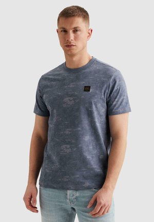 Mladý muž v modrom tričku s krátkym rukávom s potlačou a svetlomodrých džínsoch stojaci pred jednofarebným sivým pozadím.