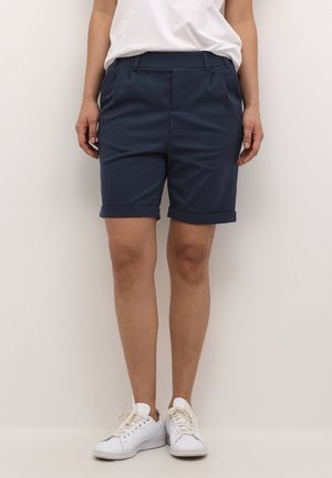 Personne portant un short bleu marine ajusté avec des revers roulés, un T-shirt blanc et des baskets blanches, debout devant un fond uni.