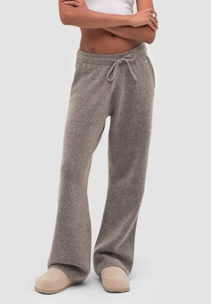 Person trägt lockere graue Strick-Sweathosen mit Kordelzug und beigen Slip-on-Schuhen, steht mit verschränkten Armen über freiem Bauch.