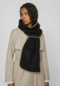Bufanda negra con una textura suave y un prominente detalle de costura dorada, usada sobre un abrigo beige con un gran cuello.