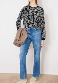 Femme portant un jean bleu, un haut à manches longues noir à motifs floraux, des baskets beiges, tenant une veste brune pliée sur un fond blanc uni.