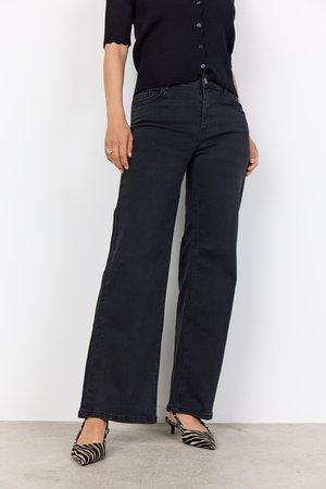 Široke črne jeans hlače z rahlo teksturo in sprednjimi žepi, v kombinaciji s črnobelim prugastim čevljem z ozkim nosom. Gumbi so vidni na srajci.