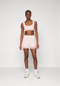 Soutien-gorge de sport rose clair avec un détail froncé à l'avant, jupe courte transparente assortie et chaussures de sport claires, le tout sur un fond uni.