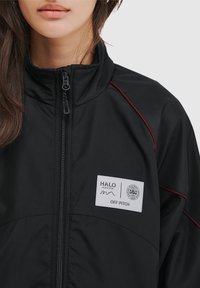 Sort jakke med høj krave, lynlås foran, røde detaljer og logo-patch. Glat tekstur, letvægtsdesign, velegnet til afslappet brug.