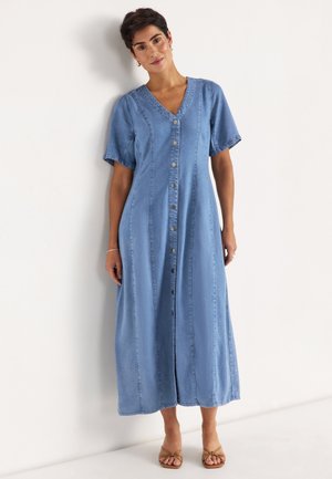 Kvinde med kort mørkt hår iført en knappet, kortærmet, ankellang blå denimkjole og beige åbne tå-sandaler, stående op ad en hvid væg.