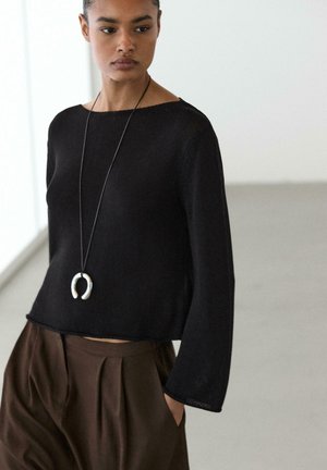 Ung kvinde iført sort langærmet top, brune højtaljede plisserede bukser og en lang halskæde med en sølv hestesko vedhæng.