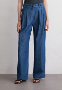 Denim wide-leg trousers in dark blue, featuring a high waist, pleated front, and relaxed fit. Worn with black minimalist sandals.