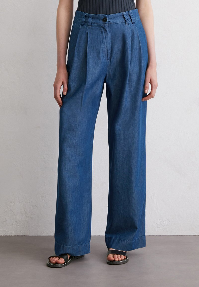 Denim wide-leg trousers in dark blue, featuring a high waist, pleated front, and relaxed fit. Worn with black minimalist sandals.