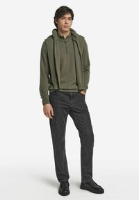 Oliwkowo zielony pulower z zamkiem przy szyi, warstwowo noszony z pasującą bluzą z kapturem, zestawiony z ciemnymi dżinsami i czarnymi butami.