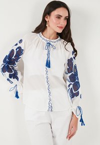 Blusa bianca con intricati ricami blu e frange sulle maniche e sul collo, con una chiusura frontale a bottoni e dettagli a pattern testurizzato.