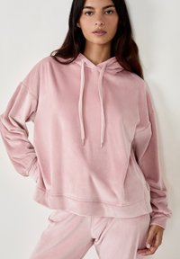 Felpa rosa, morbida, con cappuccio, dal taglio comodo, con cordini e orlo a costine. Texture soffice. Modello che indossa pantaloni abbinati.